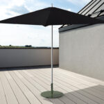 Un parasol noir est posé sur une base circulaire verte sur un toit-terrasse avec un sol en bois clair et des vues partielles du ciel et d'un paysage lointain.