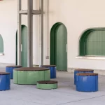A group of circular, blue and green metal stools and tables is arranged on a paved area in front of a cream-colored building with arched green windows and doors.