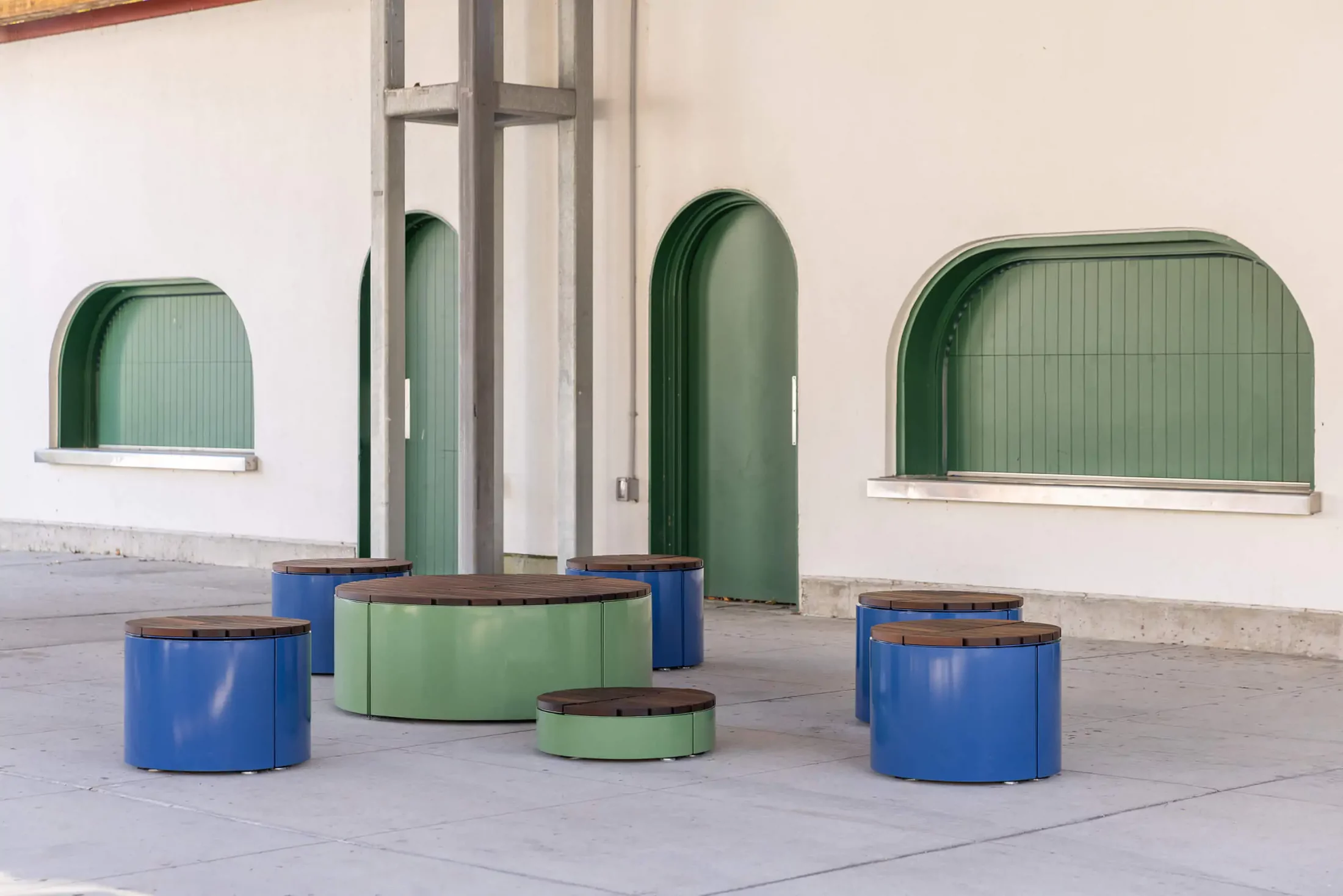 A group of circular, blue and green metal stools and tables is arranged on a paved area in front of a cream-colored building with arched green windows and doors.
