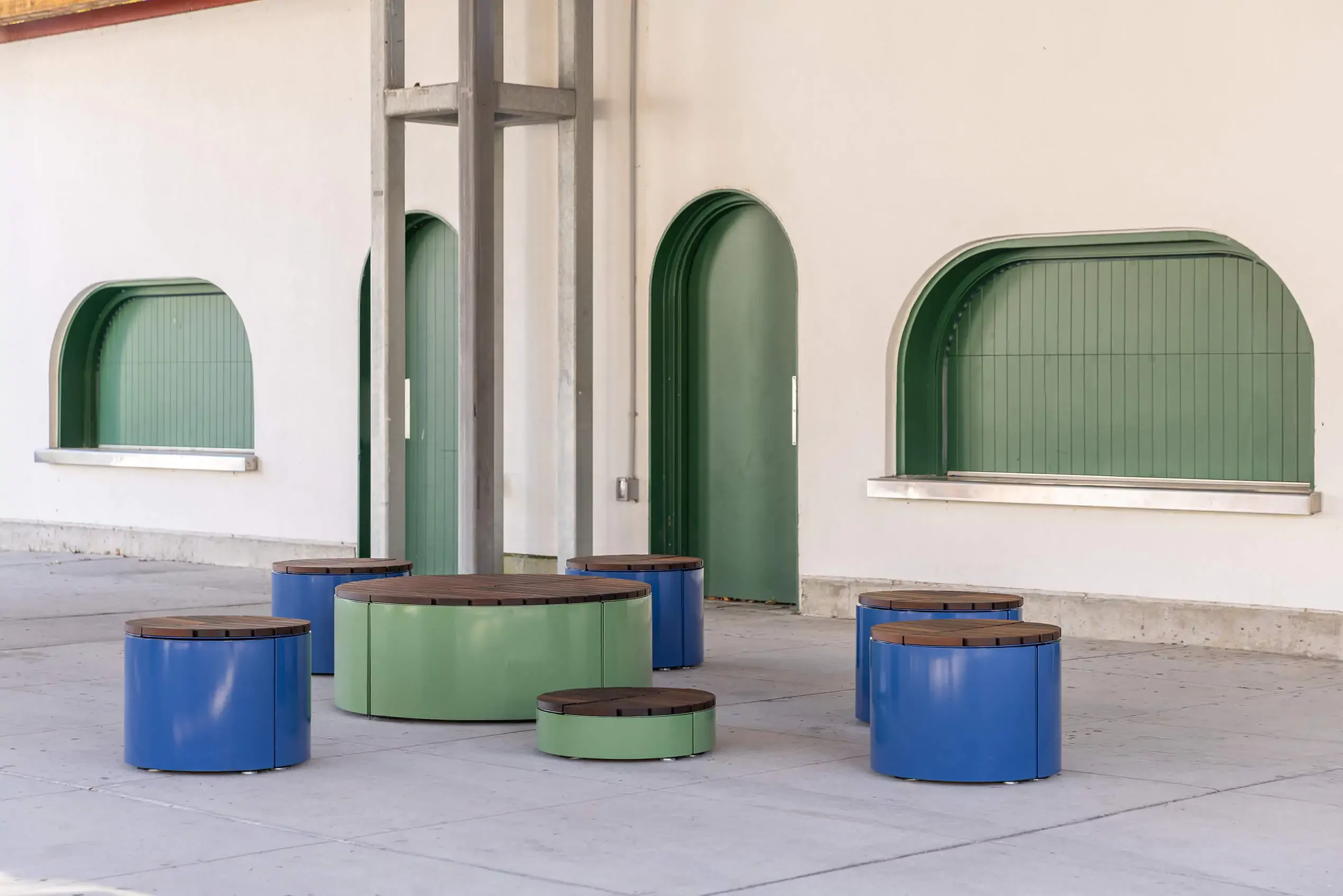 A group of circular, blue and green metal stools and tables is arranged on a paved area in front of a cream-colored building with arched green windows and doors.