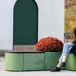 A woman in a black coat sits on a green outdoor bench beside a round potted plant, near a white building with green doors and autumn trees in the background.