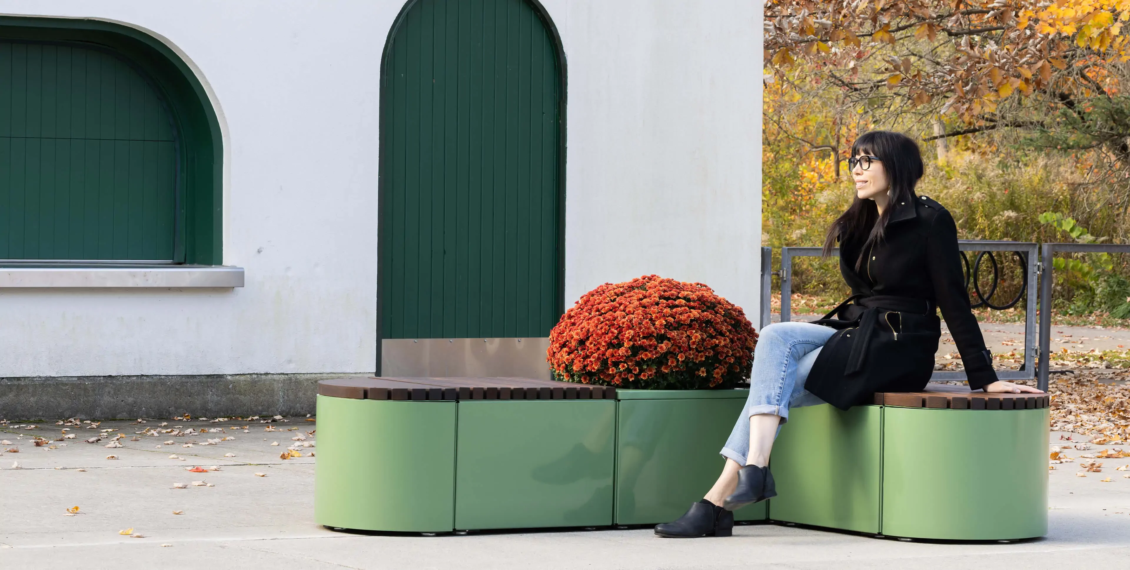 A woman in a black coat sits on a green outdoor bench beside a round potted plant, near a white building with green doors and autumn trees in the background.