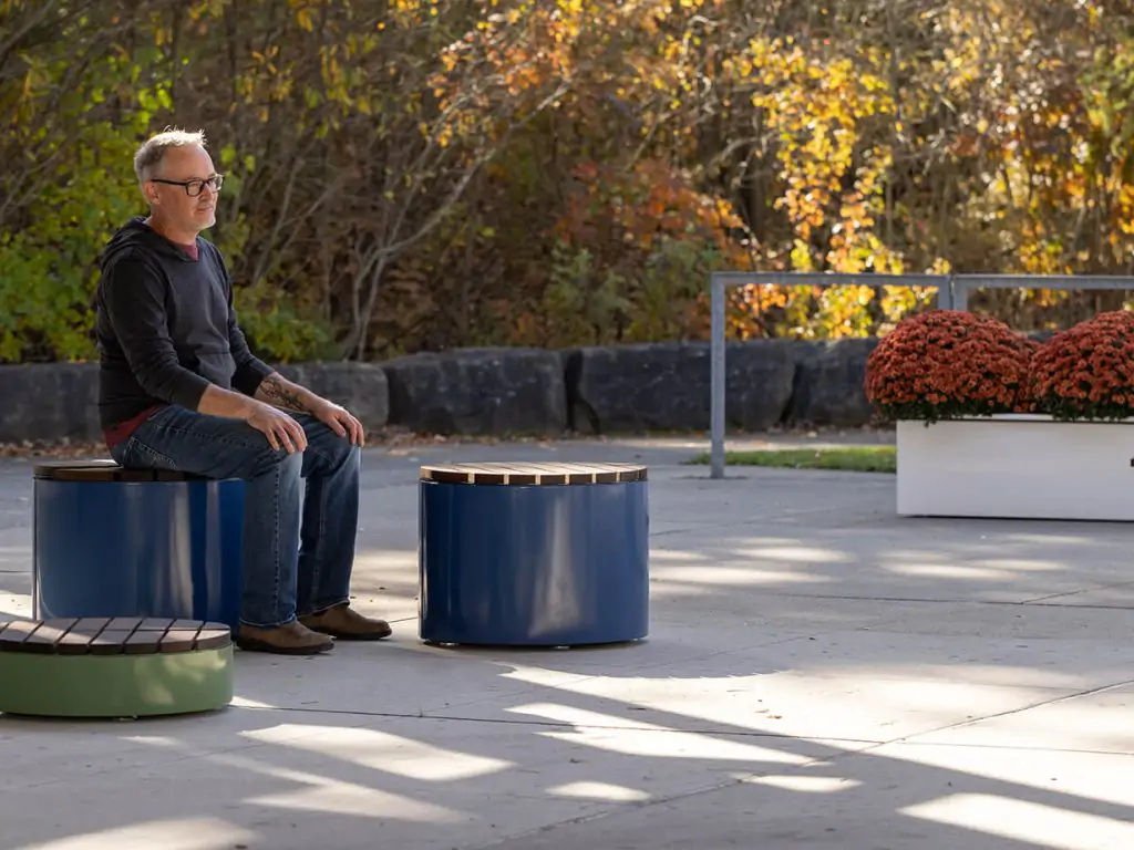 A man wearing glasses sits on a round blue bench in an outdoor area with autumn trees and potted orange chrysanthemums in the background.