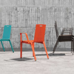 Three metal chairs—one turquoise, one orange with armrests, and a stack of four black chairs—are placed on a concrete surface in front of a textured concrete wall.