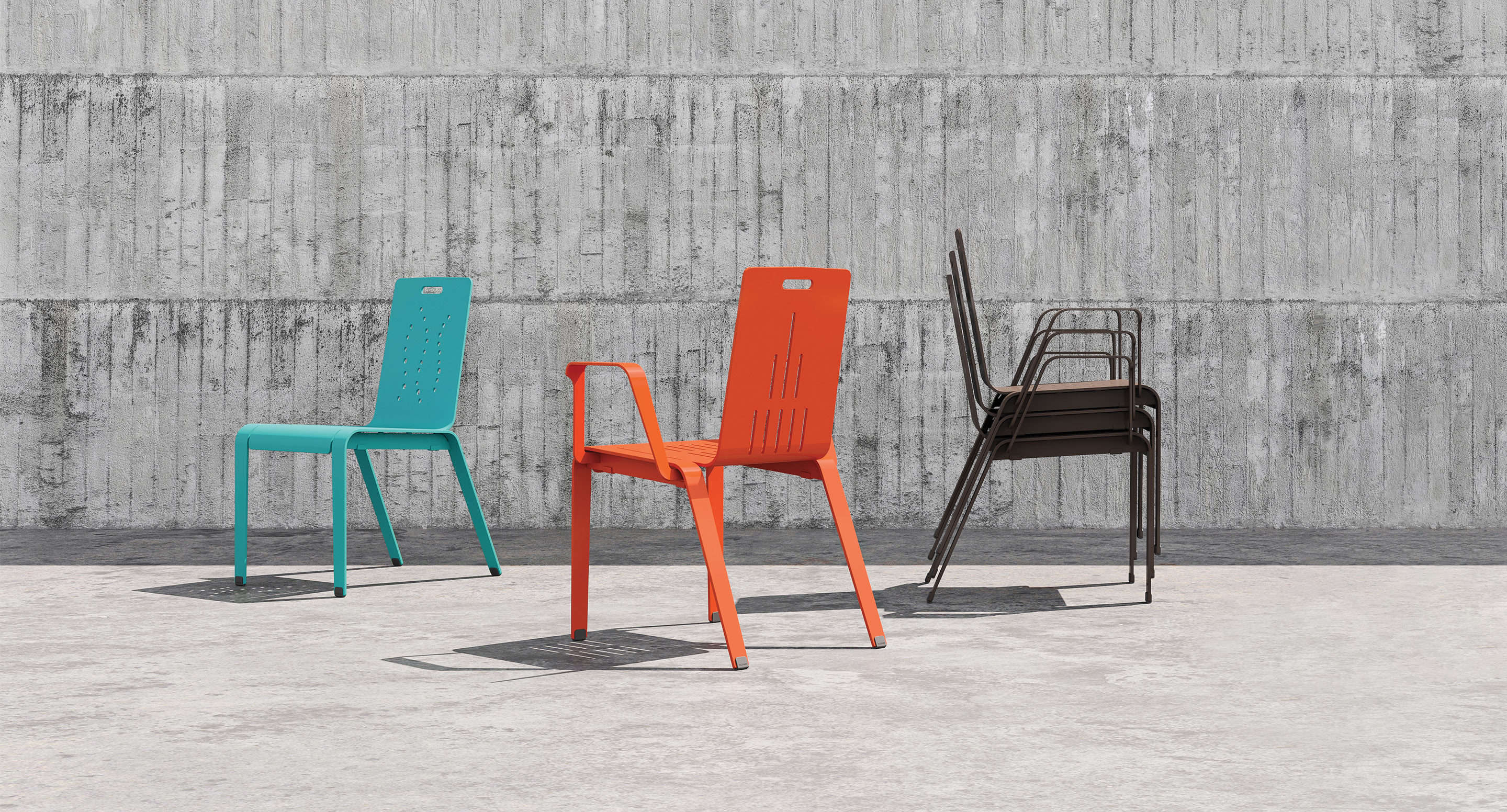 Three metal chairs—one turquoise, one orange with armrests, and a stack of four black chairs—are placed on a concrete surface in front of a textured concrete wall.