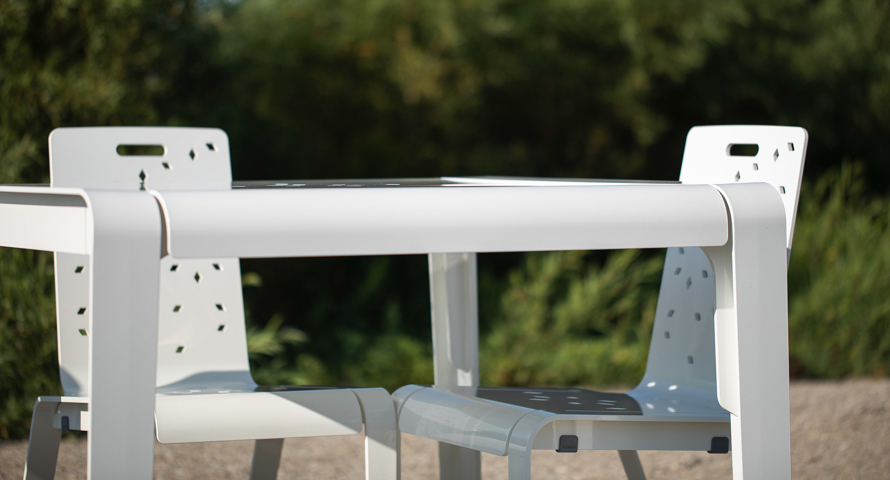 A white outdoor table with two matching white chairs positioned on a gravel surface, with green foliage in the background.