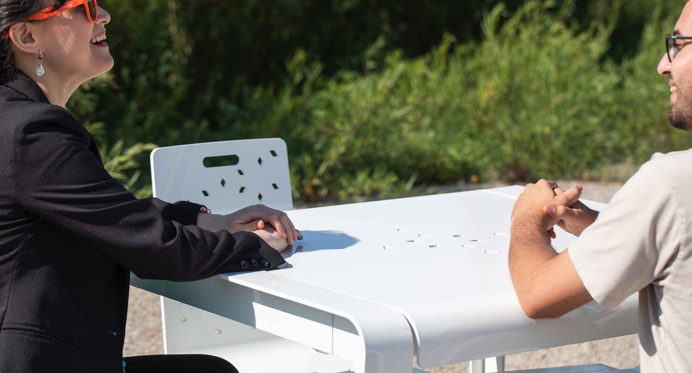 Two people sit across from each other at a white outdoor table, engaged in conversation, with greenery visible in the background.