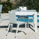 A modern white outdoor table with four matching chairs is set on a tiled patio beside a swimming pool, with plants and a stone wall in the background.
