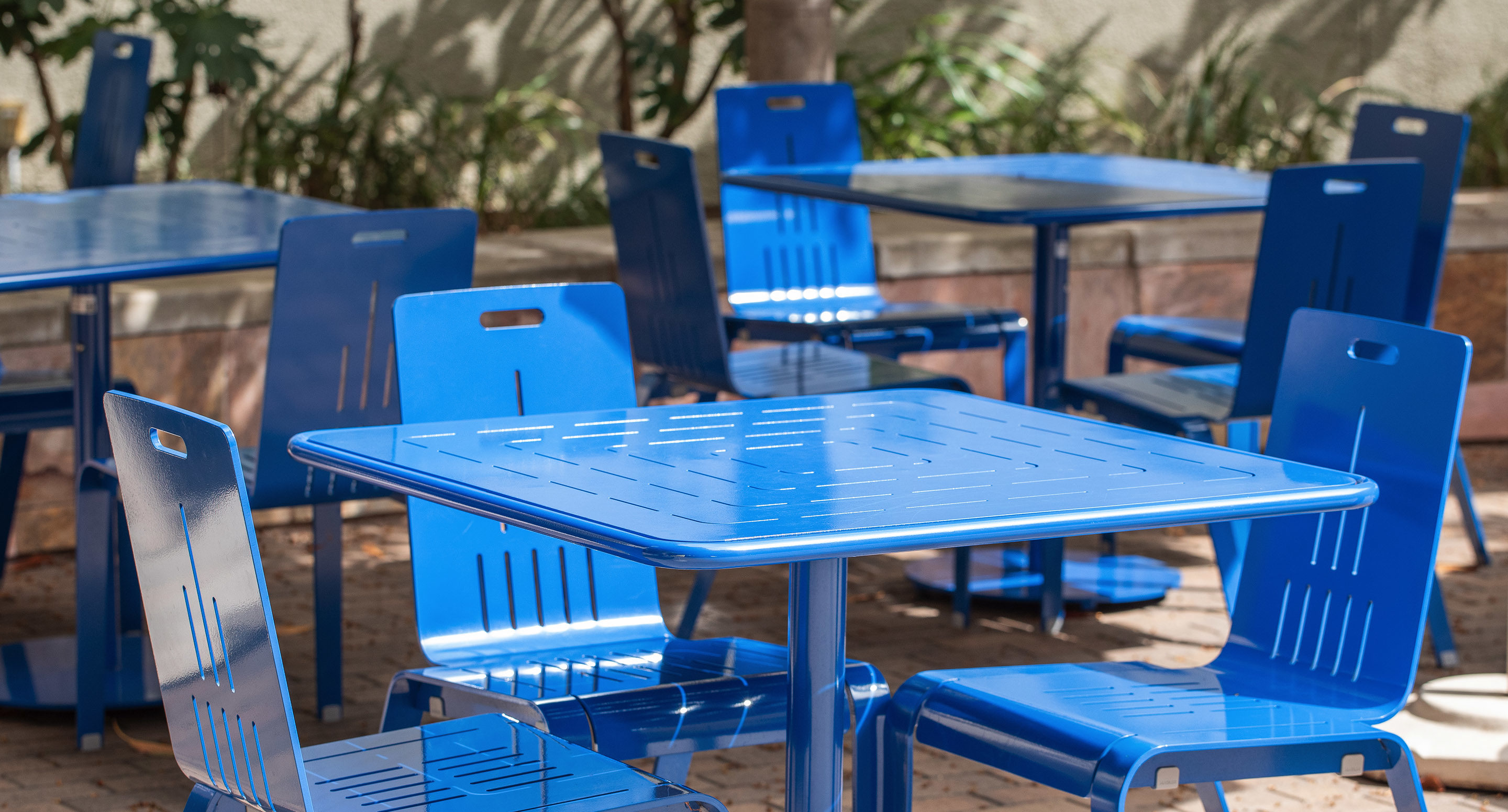Several blue metal tables and chairs are arranged outdoors on a paved surface, with greenery and a wall visible in the background.