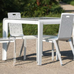 A white metal table with three matching chairs stands on a gravel path outdoors, surrounded by greenery.