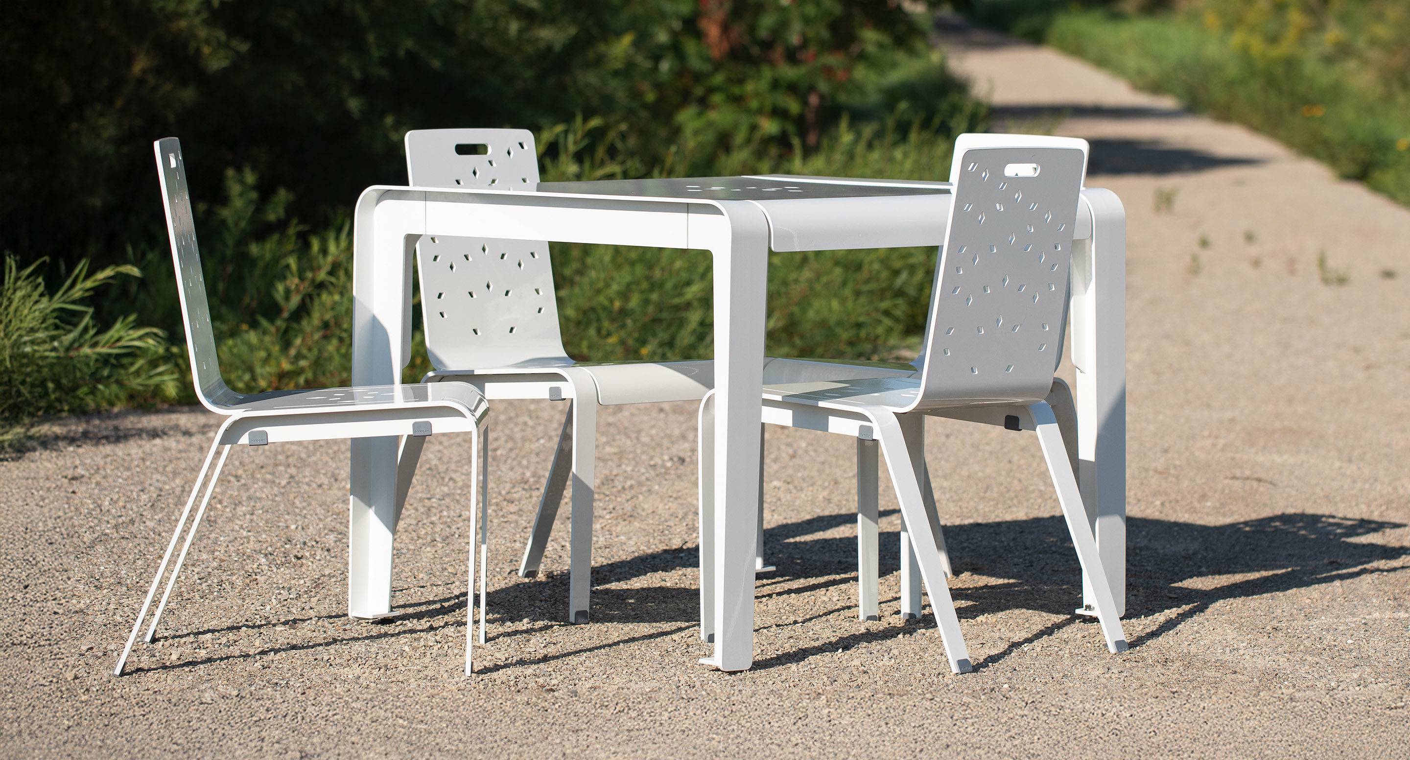 A white metal table with three matching chairs stands on a gravel path outdoors, surrounded by greenery.