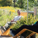 Deux personnes sont assises sur des chaises longues jaunes à l'extérieur par une journée ensoleillée, entourées de verdure et de plantes, avec une clôture et des arbres en arrière-plan.