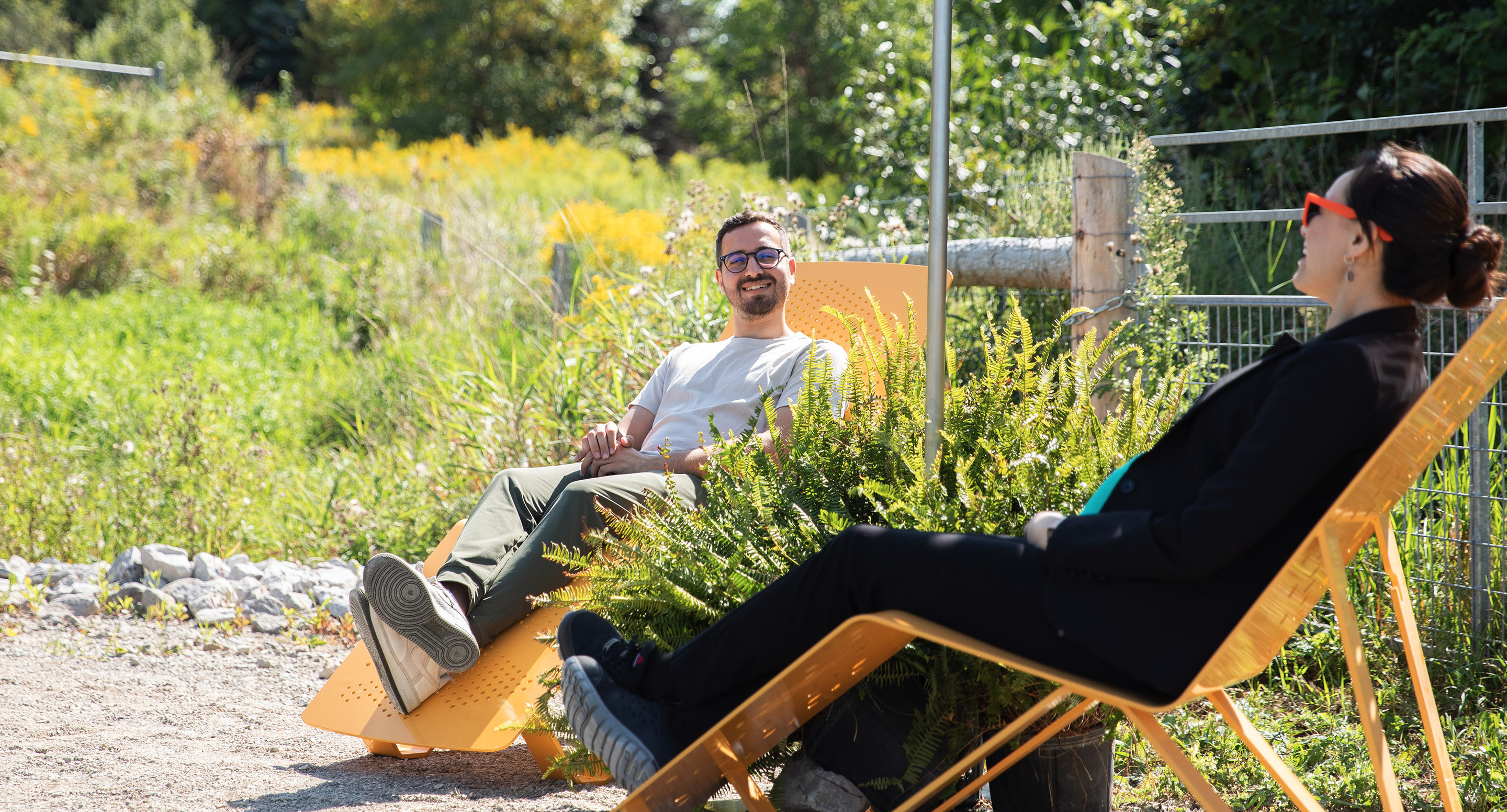 Two people sit on yellow chairs outdoors, facing each other and talking, surrounded by greenery on a sunny day.