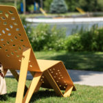 Une chaise en métal jaune avec un motif géométrique est assise sur l'herbe près d'un trottoir, avec un sac beige suspendu sur le côté et un lac en arrière-plan.