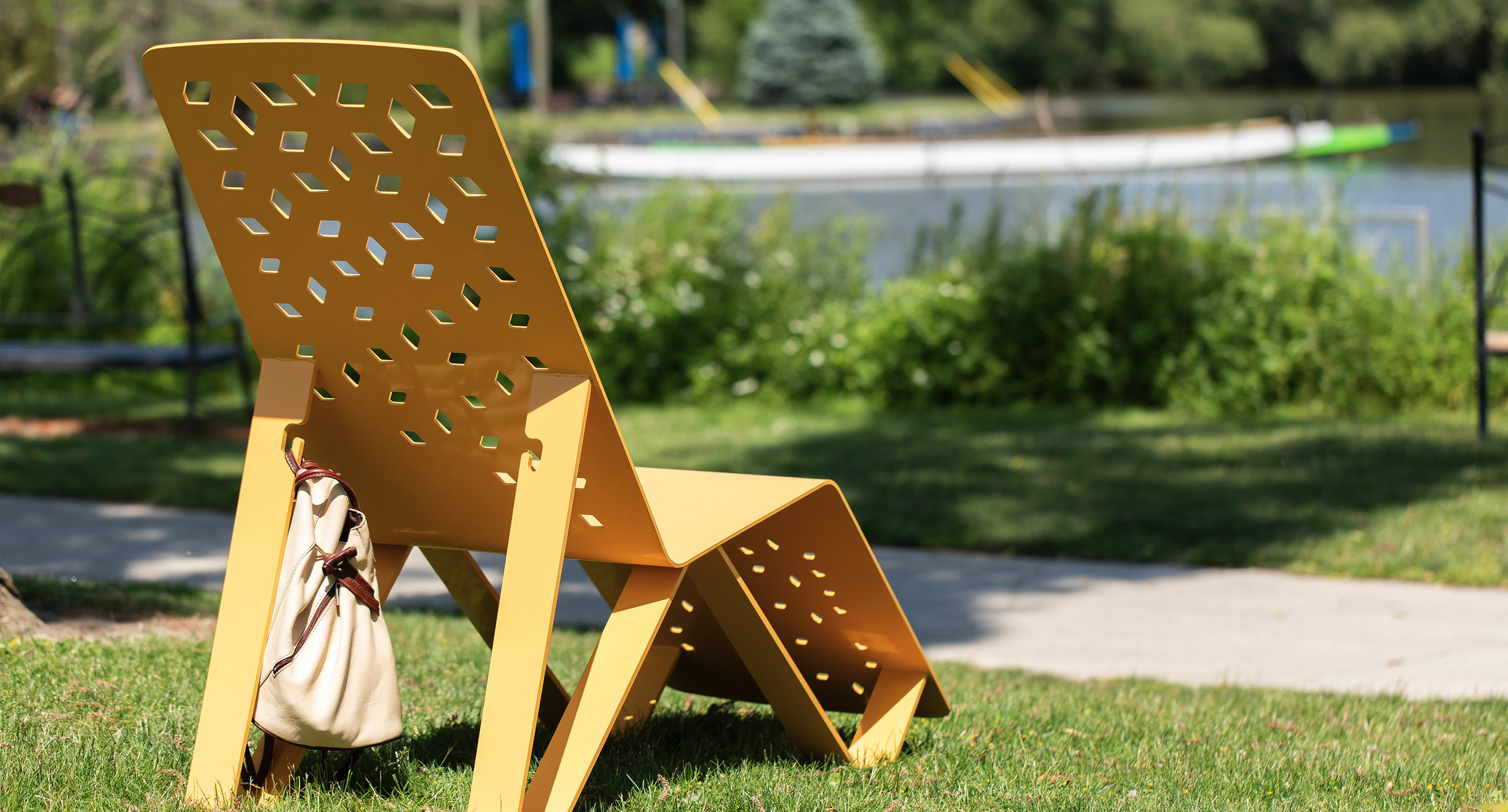 Une chaise en métal jaune avec un motif géométrique est assise sur l'herbe près d'un trottoir, avec un sac beige suspendu sur le côté et un lac en arrière-plan.