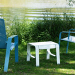 Two blue metal chairs and a small white table are placed on grass near a lake, with water and green plants in the background.