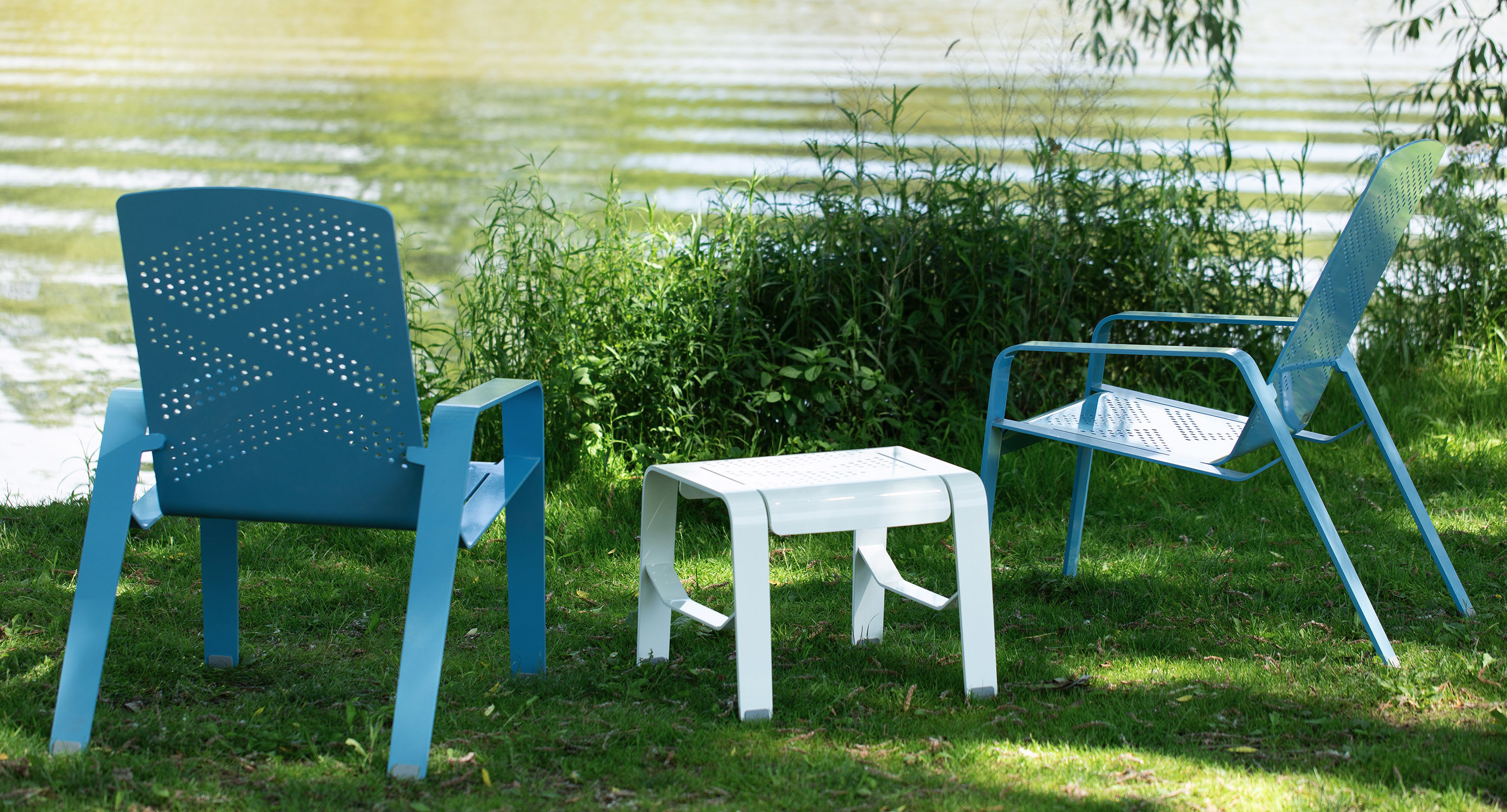 Two blue metal chairs and a small white table are placed on grass near a lake, with water and green plants in the background.