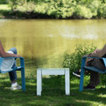 Two people sit on blue chairs facing a calm lake, with a small table between them, on a sunny day in a grassy, tree-shaded area.