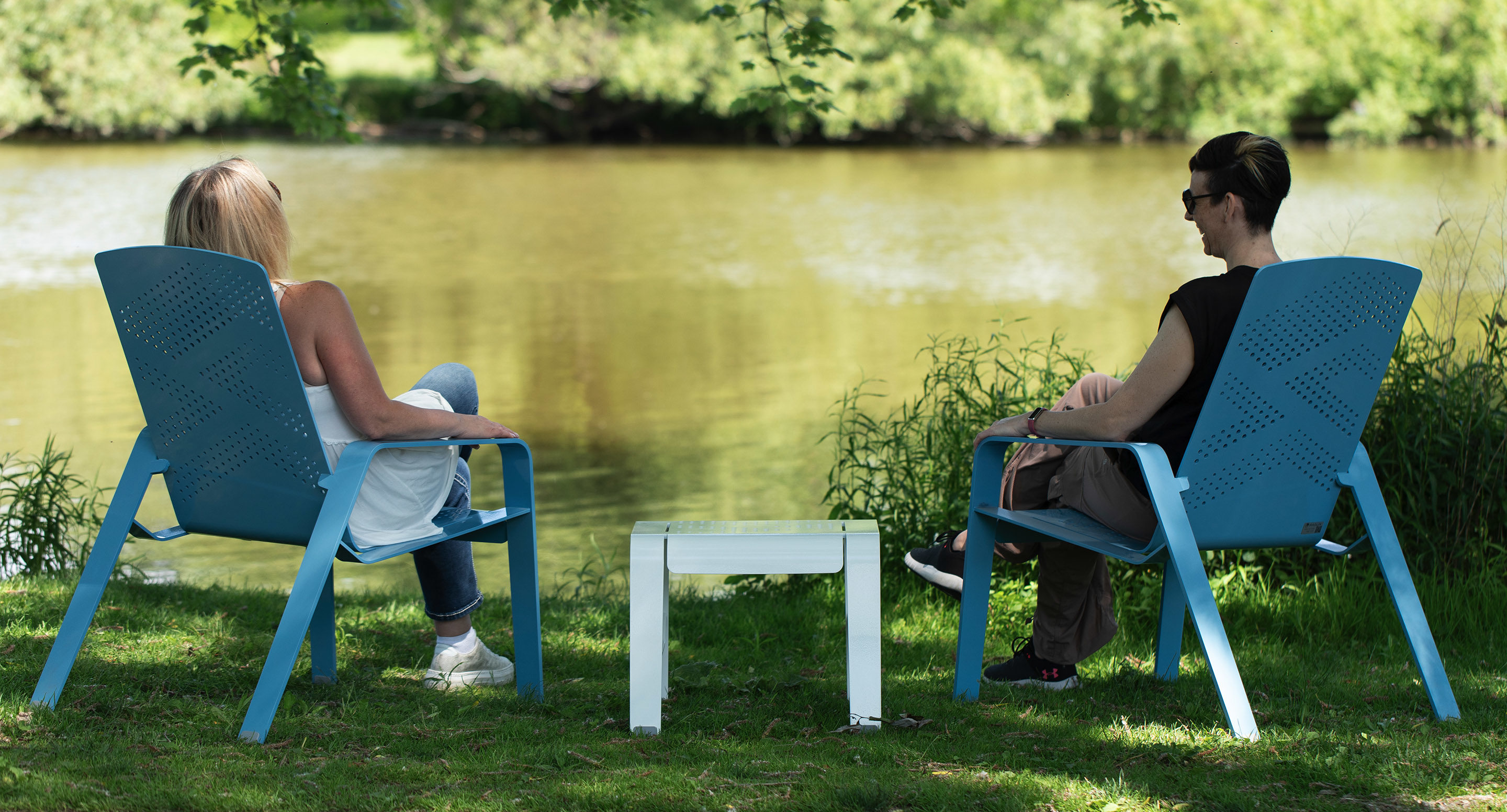 Two people sit on blue chairs facing a calm lake, with a small table between them, on a sunny day in a grassy, tree-shaded area.