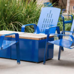 Four blue metal chairs and a rectangular blue table with a wooden top are arranged outdoors on a concrete surface, with green plants in the background.