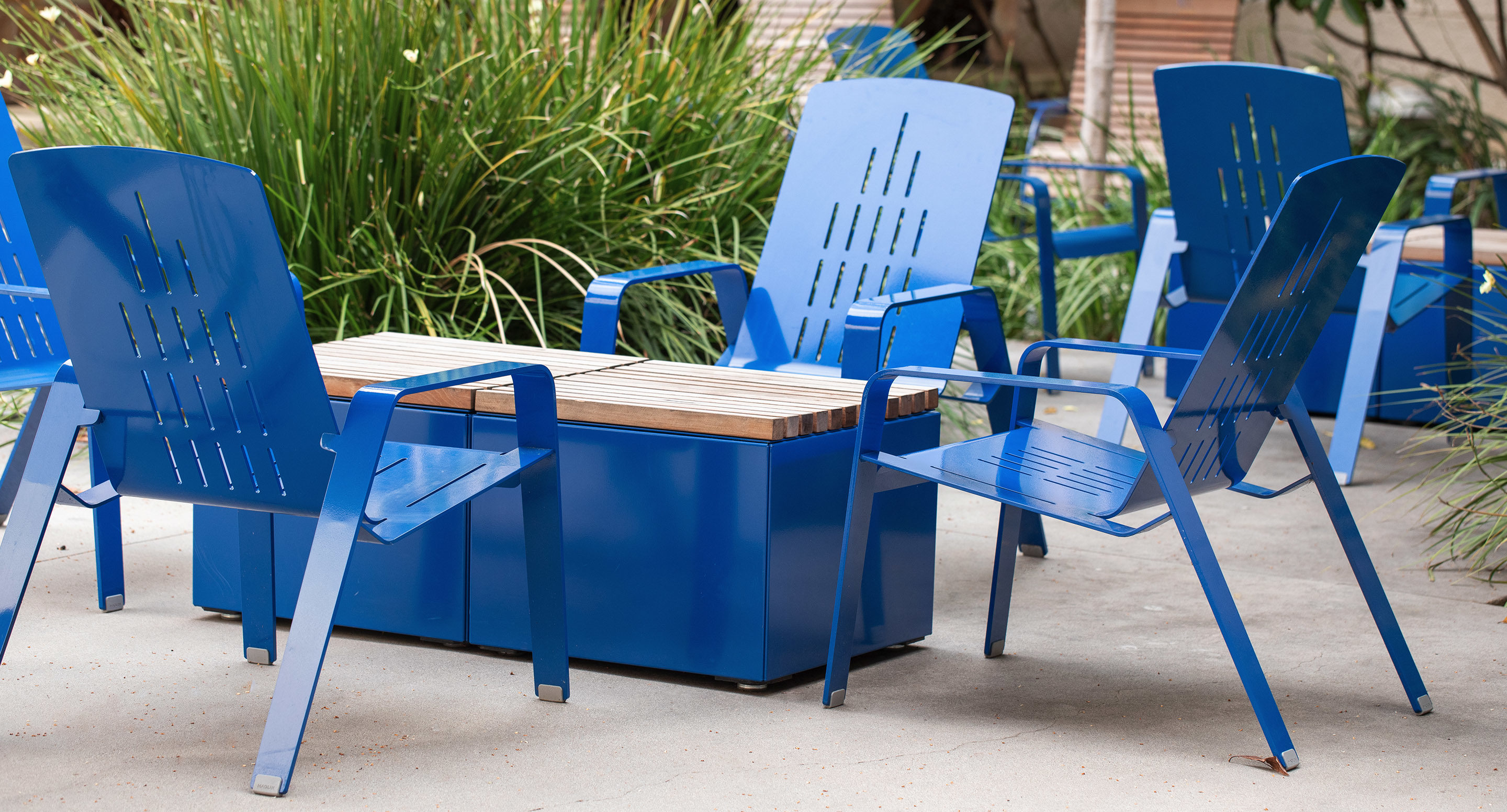 Four blue metal chairs and a rectangular blue table with a wooden top are arranged outdoors on a concrete surface, with green plants in the background.