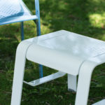 White metal stool and light blue metal chair with perforated patterns on a grassy outdoor area.