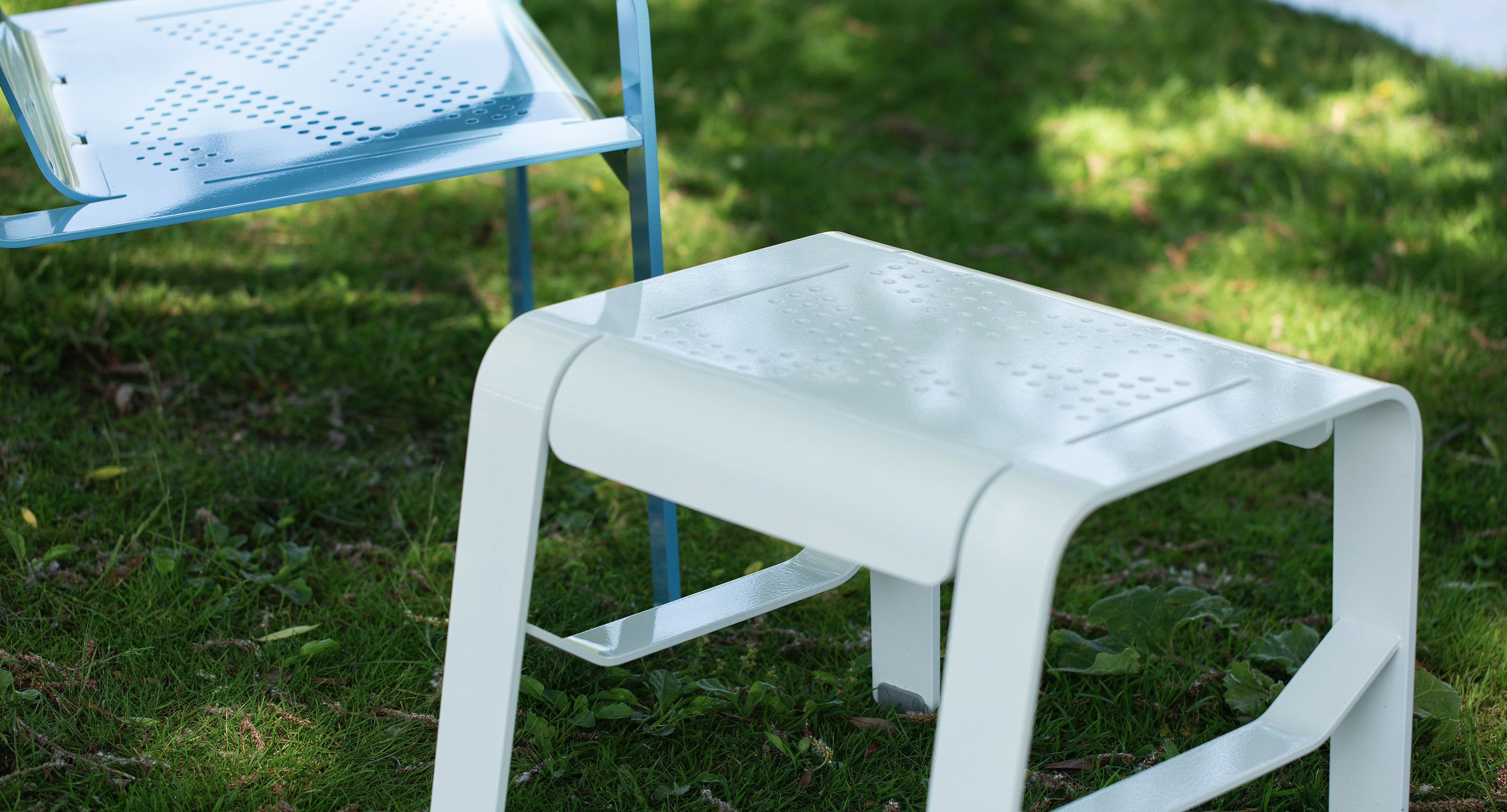 maglin_alum-lounge-table-footrest White metal stool and light blue metal chair with perforated patterns on a grassy outdoor area.
