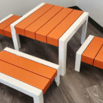 Une petite table rectangulaire aux pieds blancs et aux lattes orange, accompagnée de trois bancs assortis, sur un sol en bois.