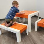 Un jeune garçon est agenouillé sur un banc orange et blanc à une table assortie, en train de dessiner ou d'écrire, dans une pièce au sol en bois clair et aux murs blancs.
