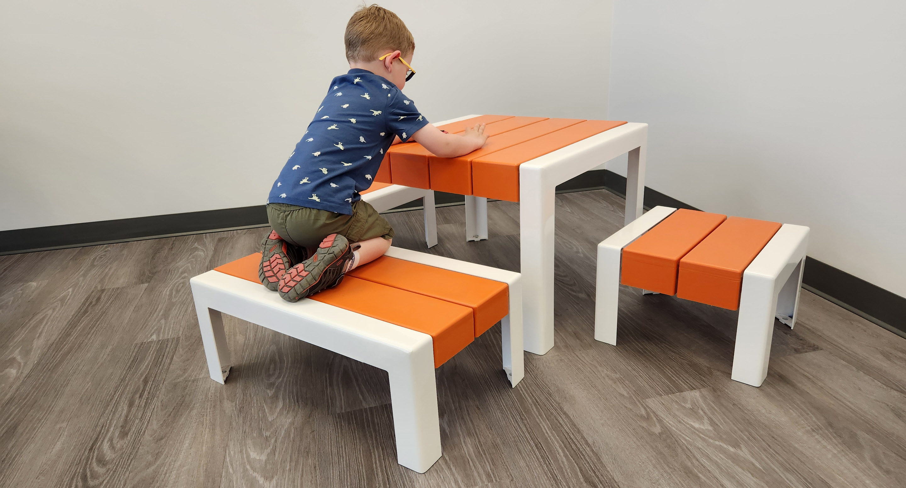 Un jeune garçon est agenouillé sur un banc orange et blanc à une table assortie, en train de dessiner ou d'écrire, dans une pièce au sol en bois clair et aux murs blancs.