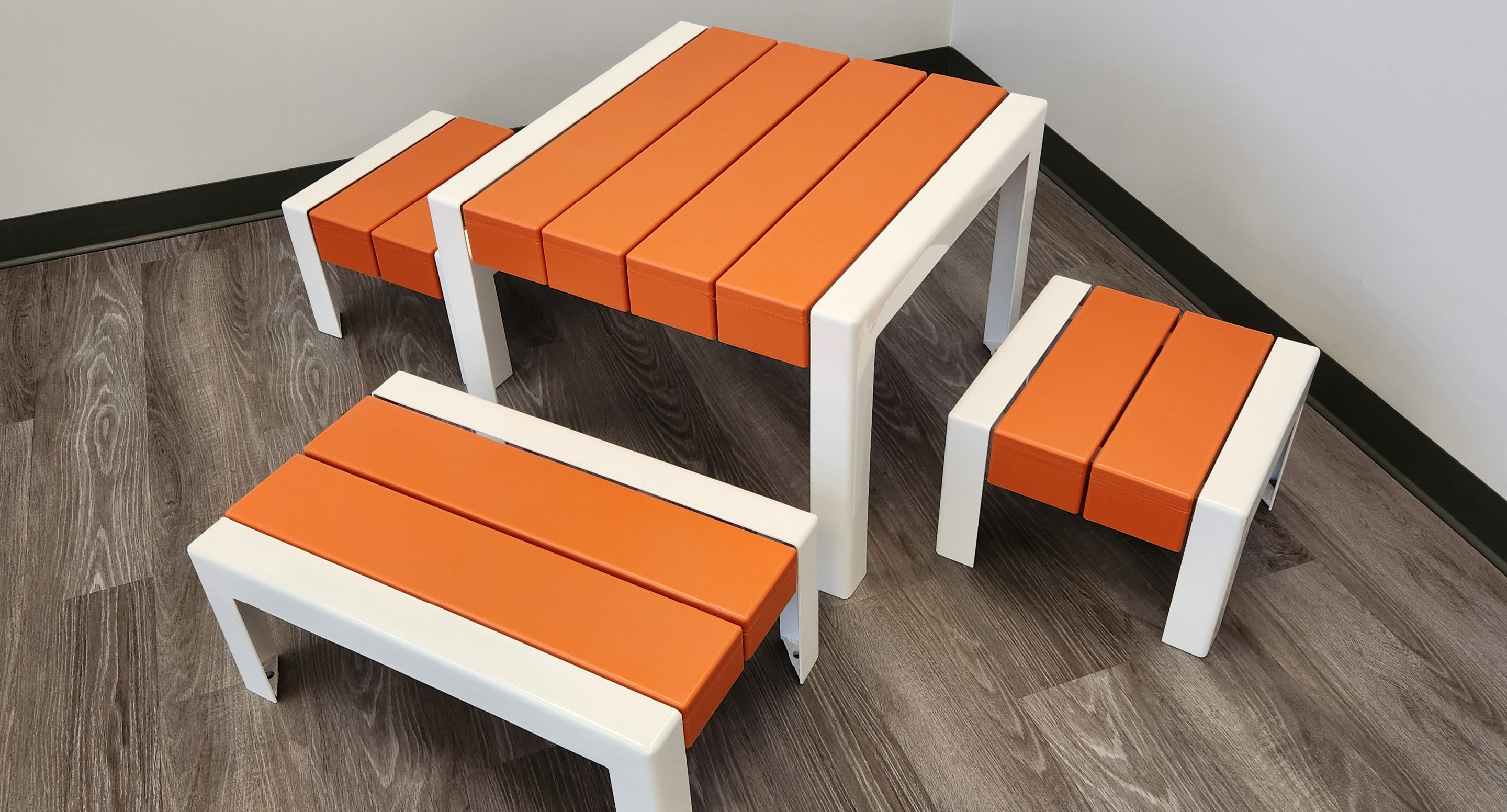 Une petite table rectangulaire aux pieds blancs et aux lattes orange, accompagnée de trois bancs assortis, sur un sol en bois.