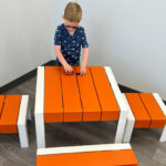 Un jeune garçon joue avec une petite voiture sur une table orange et blanche pour enfants, qui comprend deux bancs, dans une pièce aux murs gris et au sol en bois.