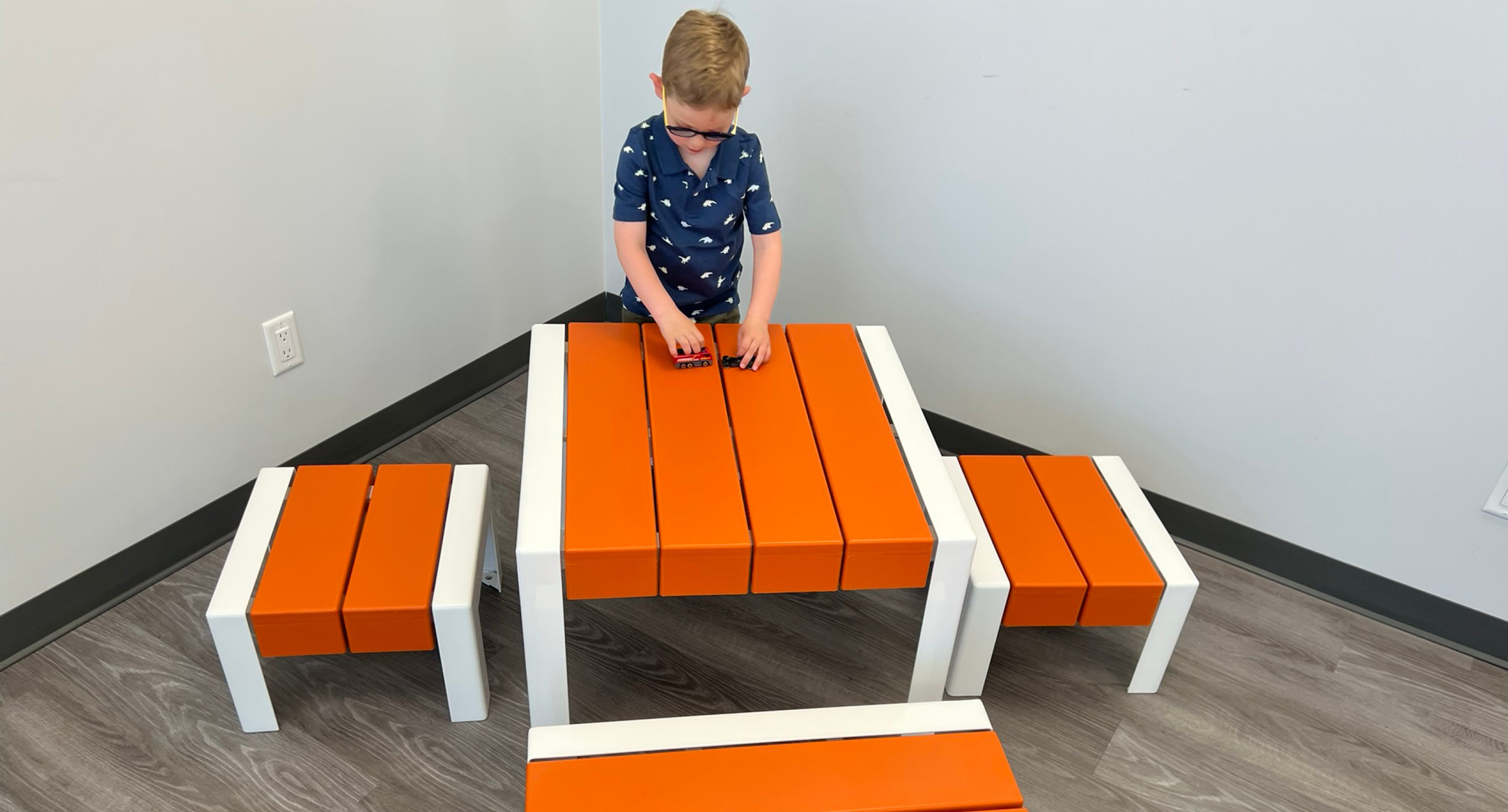 Un jeune garçon joue avec une petite voiture sur une table orange et blanche pour enfants, qui comprend deux bancs, dans une pièce aux murs gris et au sol en bois.