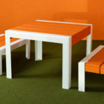 Une table blanche et deux bancs blancs avec des sièges orange sont placés sur un sol vert sur un fond orange.
