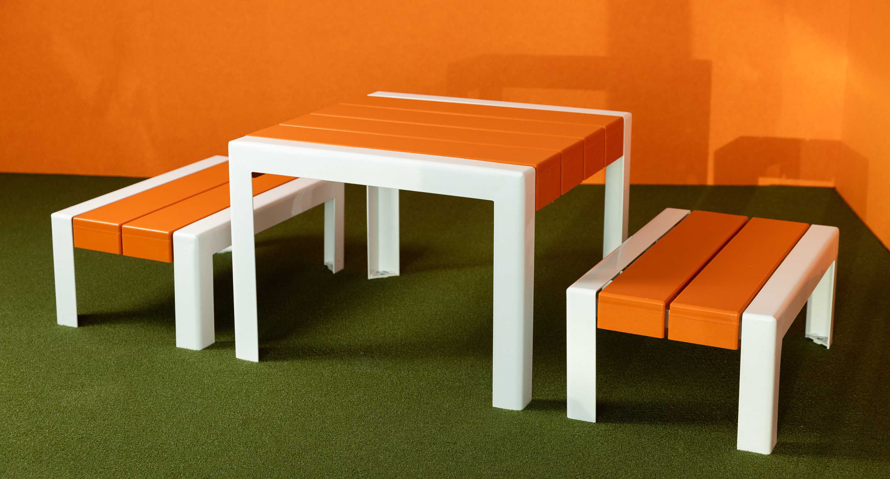 Une table blanche et deux bancs blancs avec des sièges orange sont placés sur un sol vert sur un fond orange.