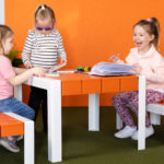 Trois jeunes filles sont assises à une table orange et blanche, lisant et coloriant des livres, dans une pièce très éclairée avec des plantes vertes et un mur orange.
