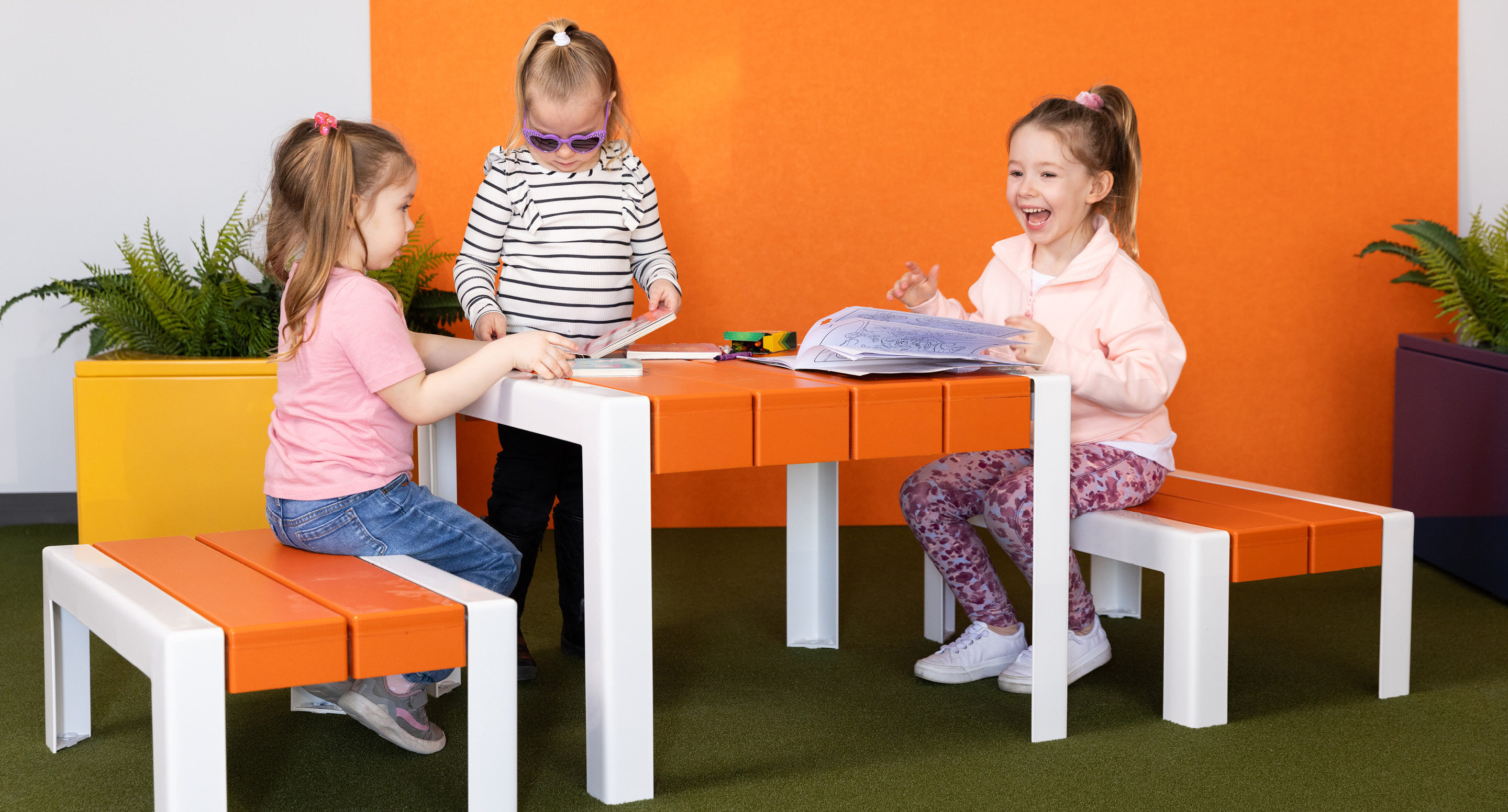 Trois jeunes filles sont assises à une table orange et blanche, lisant et coloriant des livres, dans une pièce très éclairée avec des plantes vertes et un mur orange.