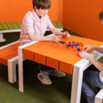 Deux garçons sont assis à une table orange et construisent avec de petits blocs en plastique dans une pièce au sol vert, aux murs orange et avec une plante dans le coin.