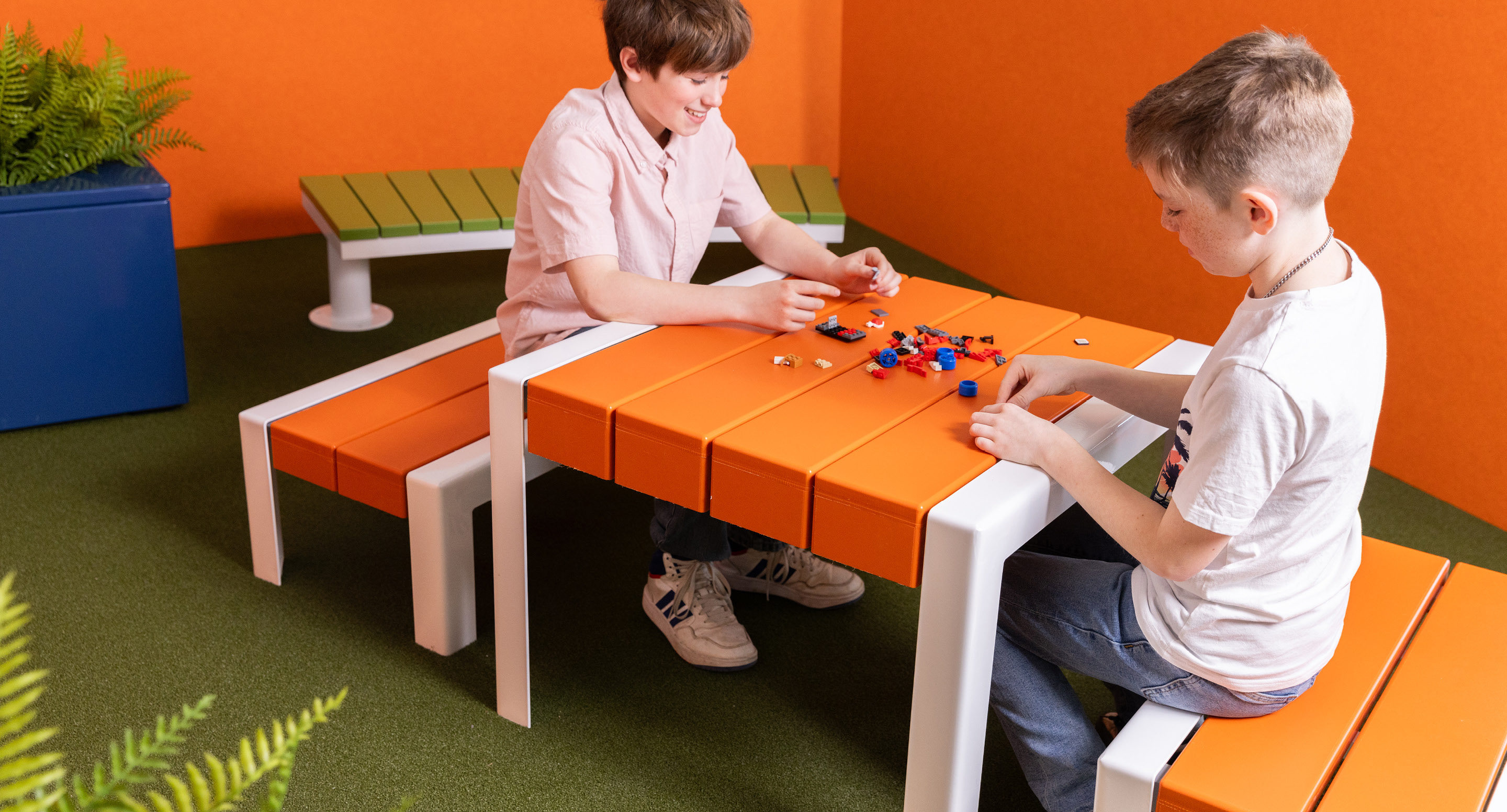 Deux garçons sont assis à une table orange et construisent avec de petits blocs en plastique dans une pièce au sol vert, aux murs orange et avec une plante dans le coin.