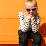Un jeune enfant portant des lunettes de soleil violettes en forme de cœur et une chemise rayée est assis sur un banc orange sur un fond orange, souriant avec les mains sous le menton.