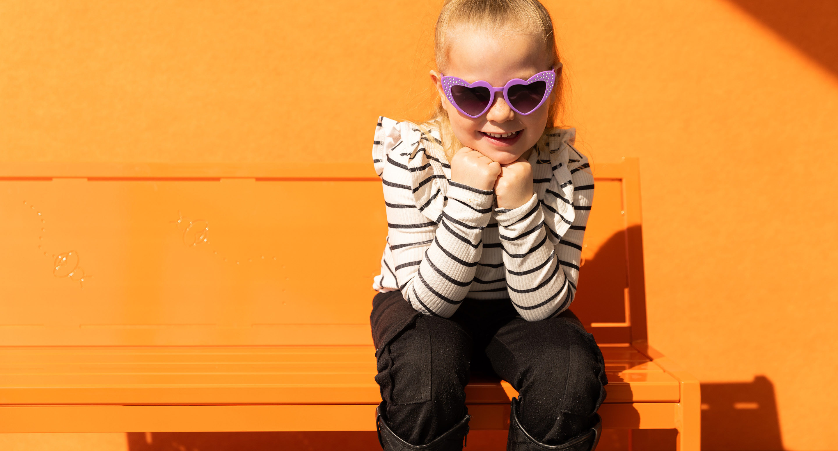 maglin_childrens-400-benches-1 Un jeune enfant portant des lunettes de soleil violettes en forme de cœur et une chemise rayée est assis sur un banc orange sur un fond orange, souriant avec les mains sous le menton.