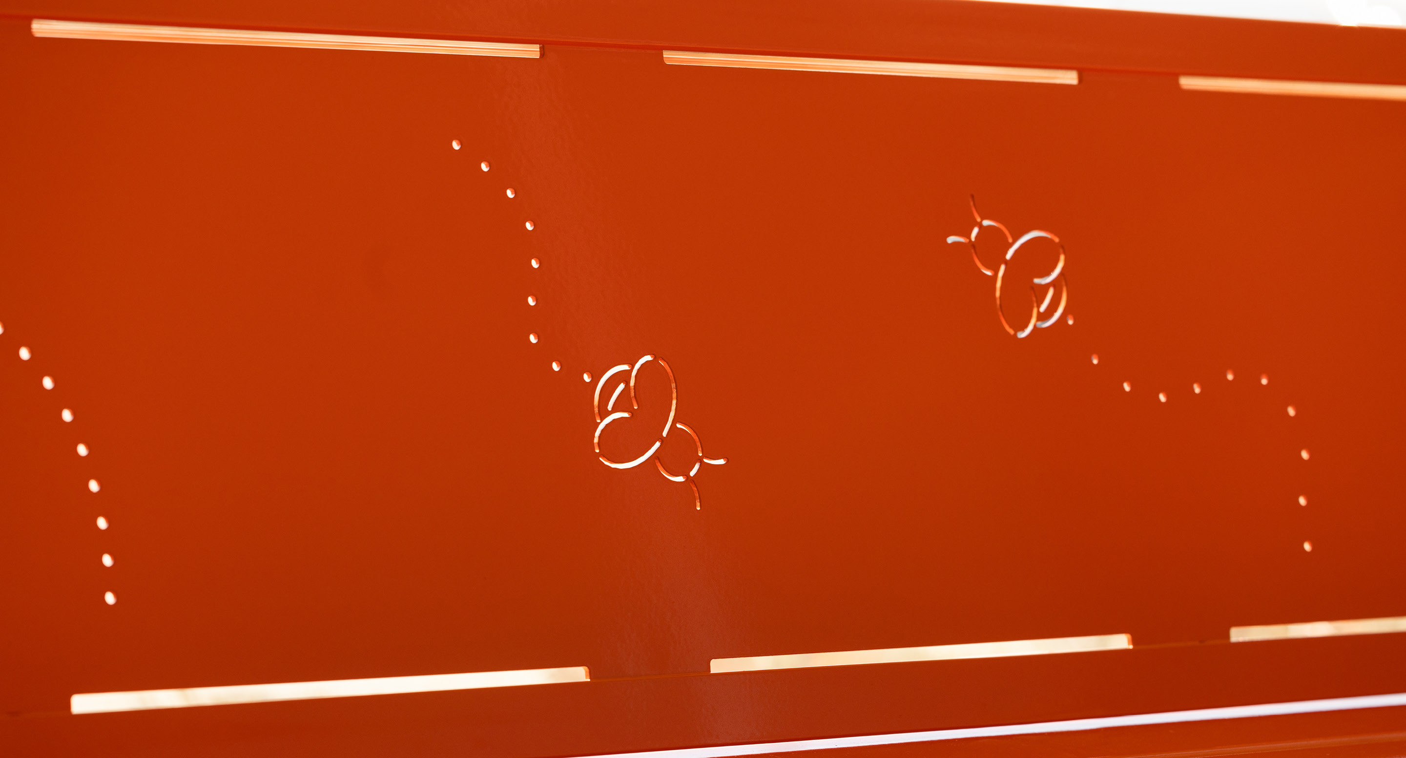maglin_childrens-400-benches-2 Surface métallique orange avec deux découpes en forme d'abeille et des lignes pointillées, permettant à la lumière de passer à travers le motif.