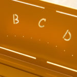 Gros plan d'un banc en métal jaune dont le dossier est orné de lettres A, B, C, D et E découpées, chacune accompagnée de points Braille correspondants sous les lettres.