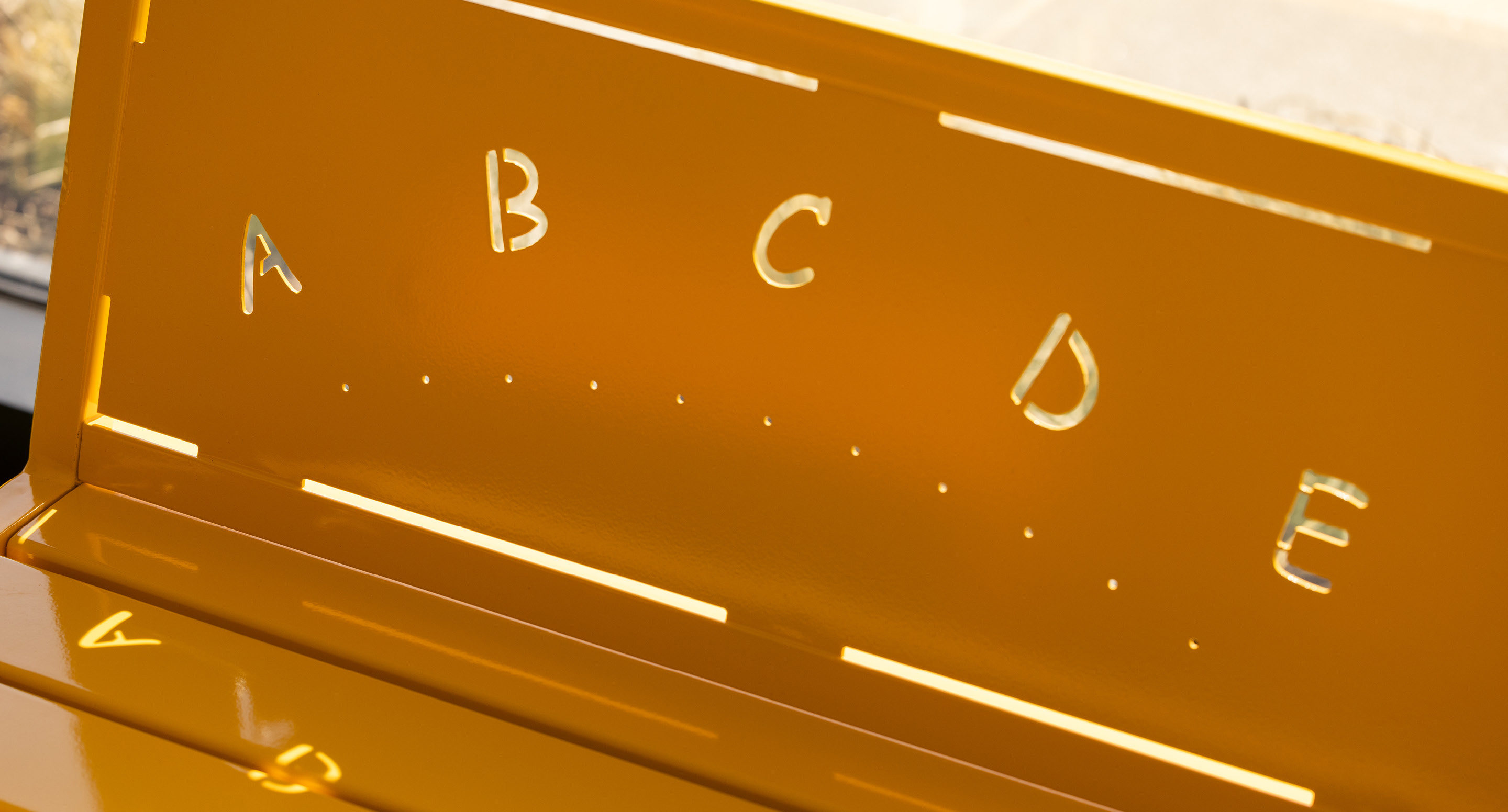 maglin_childrens-400-benches-3 Gros plan d'un banc en métal jaune dont le dossier est orné de lettres A, B, C, D et E découpées, chacune accompagnée de points Braille correspondants sous les lettres.