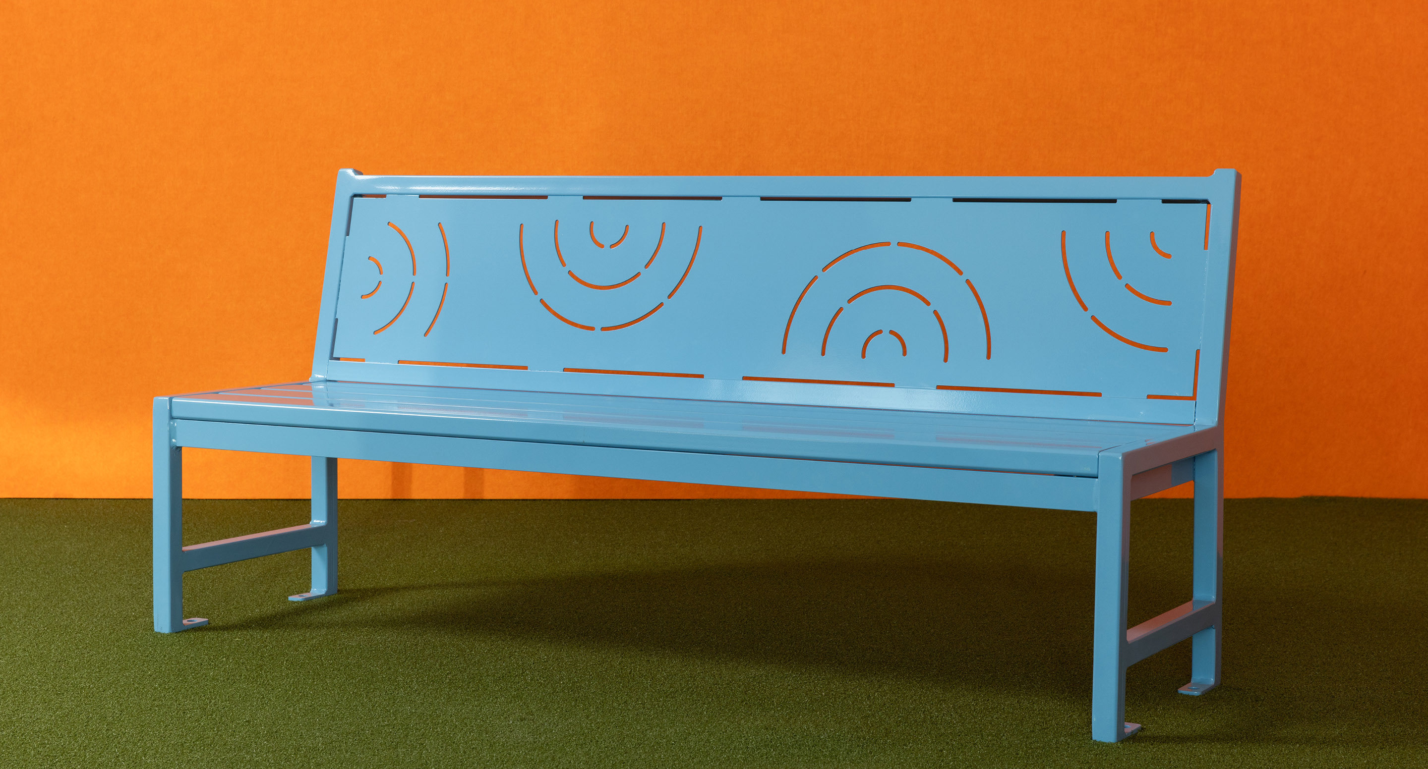 maglin_childrens-400-benches-4 Un banc métallique bleu avec des motifs circulaires est posé sur un gazon artificiel vert contre un mur orange.