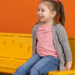 Une jeune fille avec une queue de cheval est assise sur un banc jaune avec des lettres de l'alphabet, en souriant, sur un fond orange et gris.
