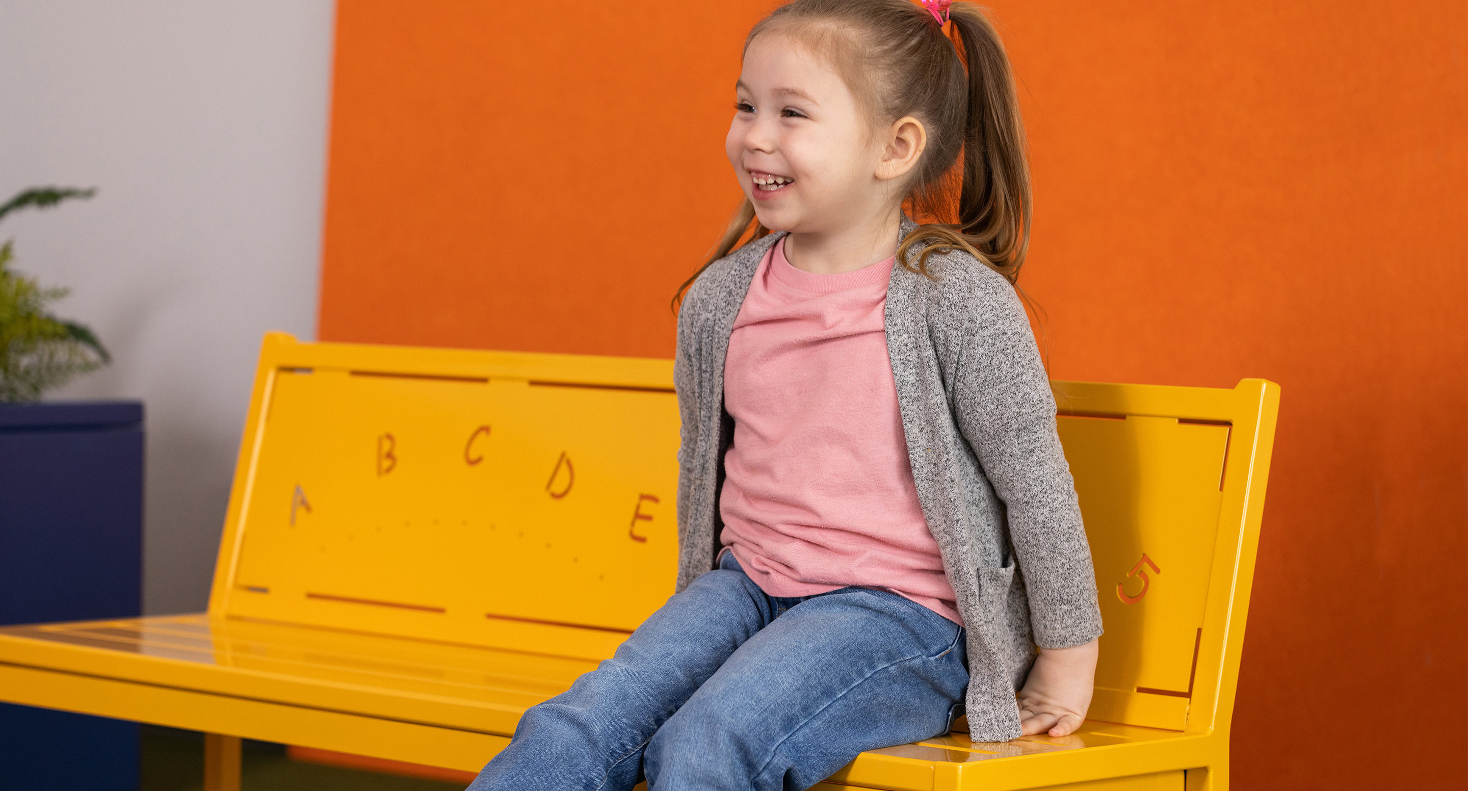 maglin_childrens-400-benches-5 Une jeune fille avec une queue de cheval est assise sur un banc jaune avec des lettres de l'alphabet, en souriant, sur un fond orange et gris.