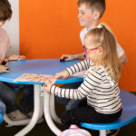 Quatre enfants sont assis autour d'une table bleue, s'occupant d'une feuille d'activité colorée et de petits objets, contre un mur orange.