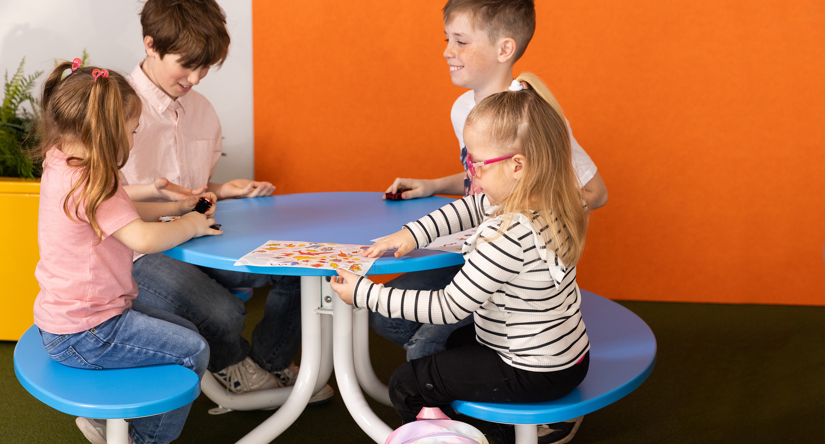 maglin_childrens-fava-picnic-tables-1 Quatre enfants sont assis autour d'une table bleue, s'occupant d'une feuille d'activité colorée et de petits objets, contre un mur orange.