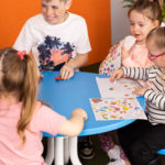 Quatre enfants sont assis autour d'une table bleue, s'amusant avec des autocollants et du papier, tandis qu'un autre enfant se tient à proximité. Il semble s'agir d'une salle de classe ou d'une aire de jeux.